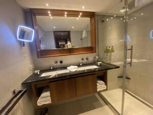 The spacious bathroom of a luxury cabin on a cruise ship with double sinks and a glass walled shower.