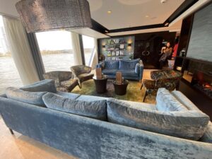 The interior living room area of a cruise ship with blue crushed velvet couch and floral patterned furniture. One wall i windows with a view of the river and land.
