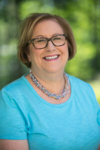 Fay deHaas smiling in a blue shirt. Sheis wearting glasses and and a colorful beaded necklace.