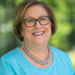 Fay deHaas smiling in a blue shirt. Sheis wearting glasses and and a colorful beaded necklace.