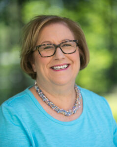 Fay deHaas smiling in a blue shirt. Sheis wearting glasses and and a colorful beaded necklace.