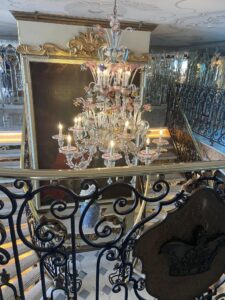 The lobby staircase of a luxury river cruise ship with a crystal chandelier and decorative wrought iron banisters along the stairs.
