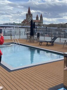The deck of a cruise ship showing a swimming pool and deck furniture and the view of a large church in Vienna in the background.