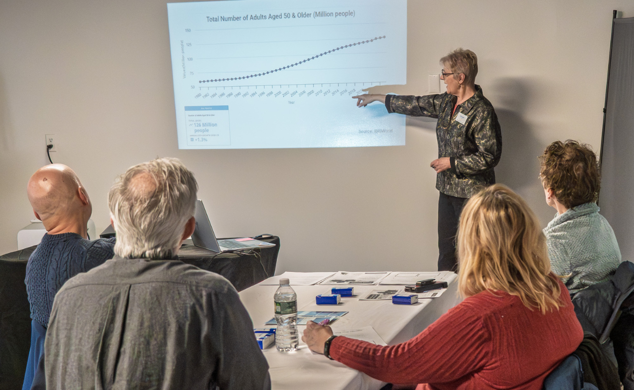 Jill Kovalich, founder of SoloAgersConnect.com leading a workshop presentation for solo agers to four attendees, pointing to a projected graph showing the total number of adults aged 50 and older over time, with charts and papers on the table in front of the participants.