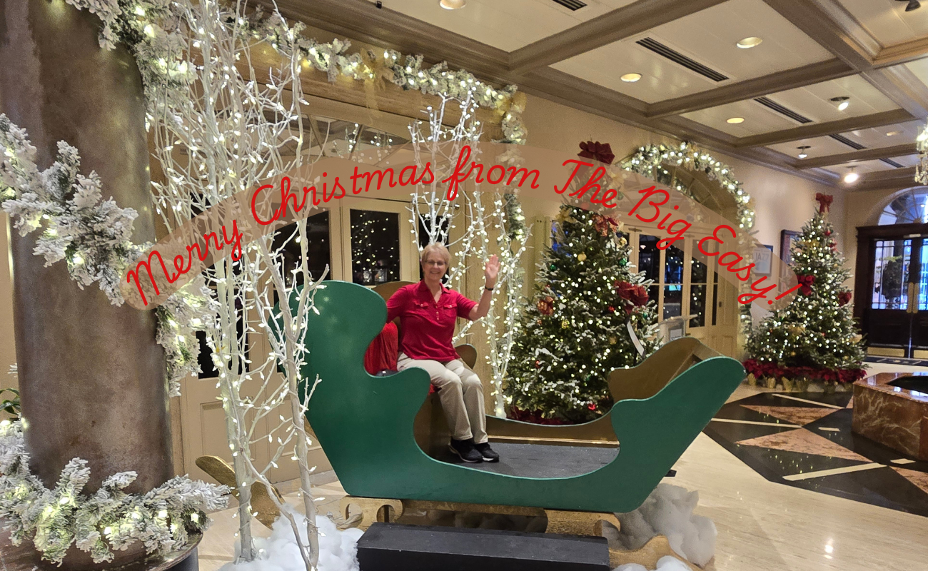 A woman smiling and waving sitting in a big sleigh in a hotel lobby decorated for Christmas with the words "Merry Christmas from The Big Easy!"