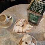 Table with coffee and a plate of three beignet donuts covered in powdered sugar.