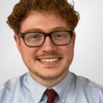 Photo of a young man with curly red hair and glasses wearing a light blue shirt and dark red tie.
