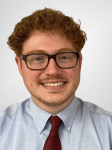 Photo of a young man with curly red hair and glasses wearing a light blue shirt and dark red tie.