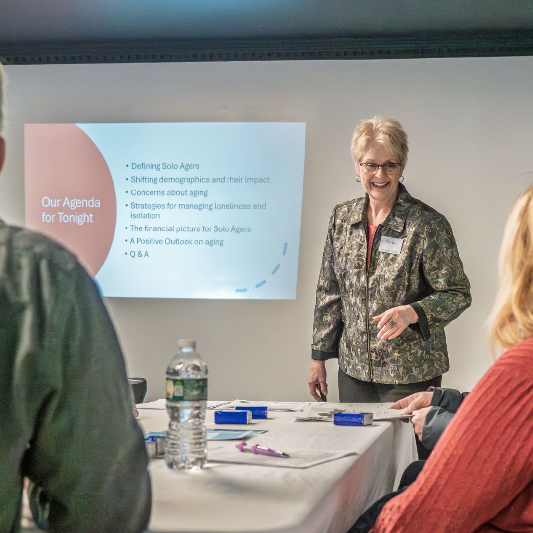Jill Kovalich presenting her interactive workshop “Solo Agers on the Rise.” A middle aged woman with blonde hair and glasses smiling as she delivers a presentation to an audience about solo Agers. On the wall behind her is a powerpoint slide with and agenda that says: Our Agenda for Tonight, Defining Solo Agers, Shifting demographics and their impact, concerns about aging, strategies for managing loneliness, the financial picture for solo agers, a positive outlook on aging, and Q&A.