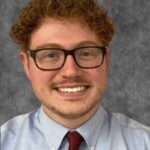 Photo of a young man with curly red hair and glasses wearing a light blue shirt and dark red tie.